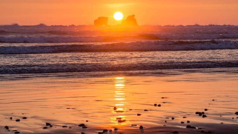 Warme, rote Lichtstimmung &uuml;ber Strand und Meer, am Horzont ein gelben Feuerballen, der zwischen zwei Felsen versinkt